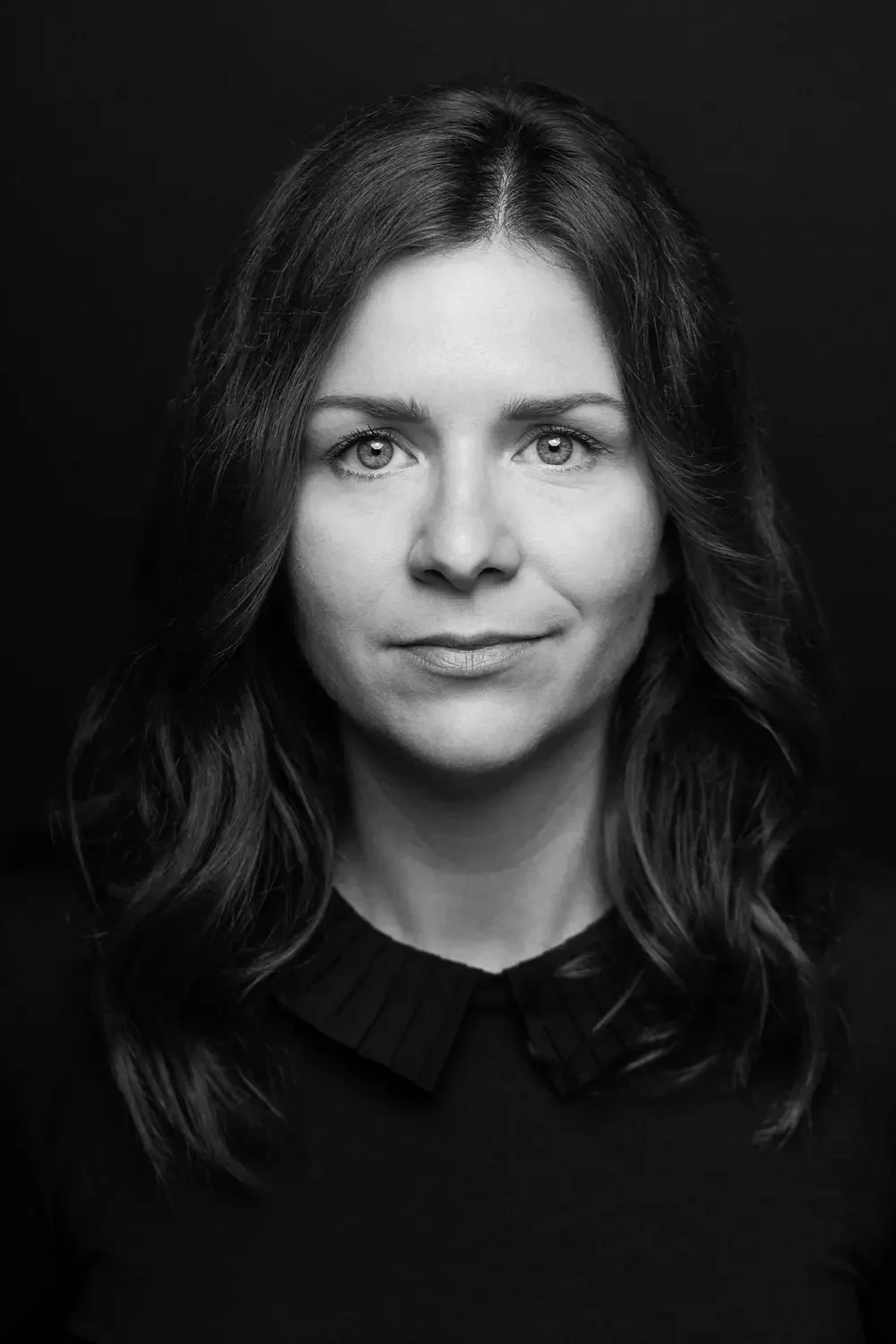 Schwarz-Weiß-Porträt einer Frau mit dunklen, gewellten Haaren und direktem Blick, gegen schwarzen Hintergrund.