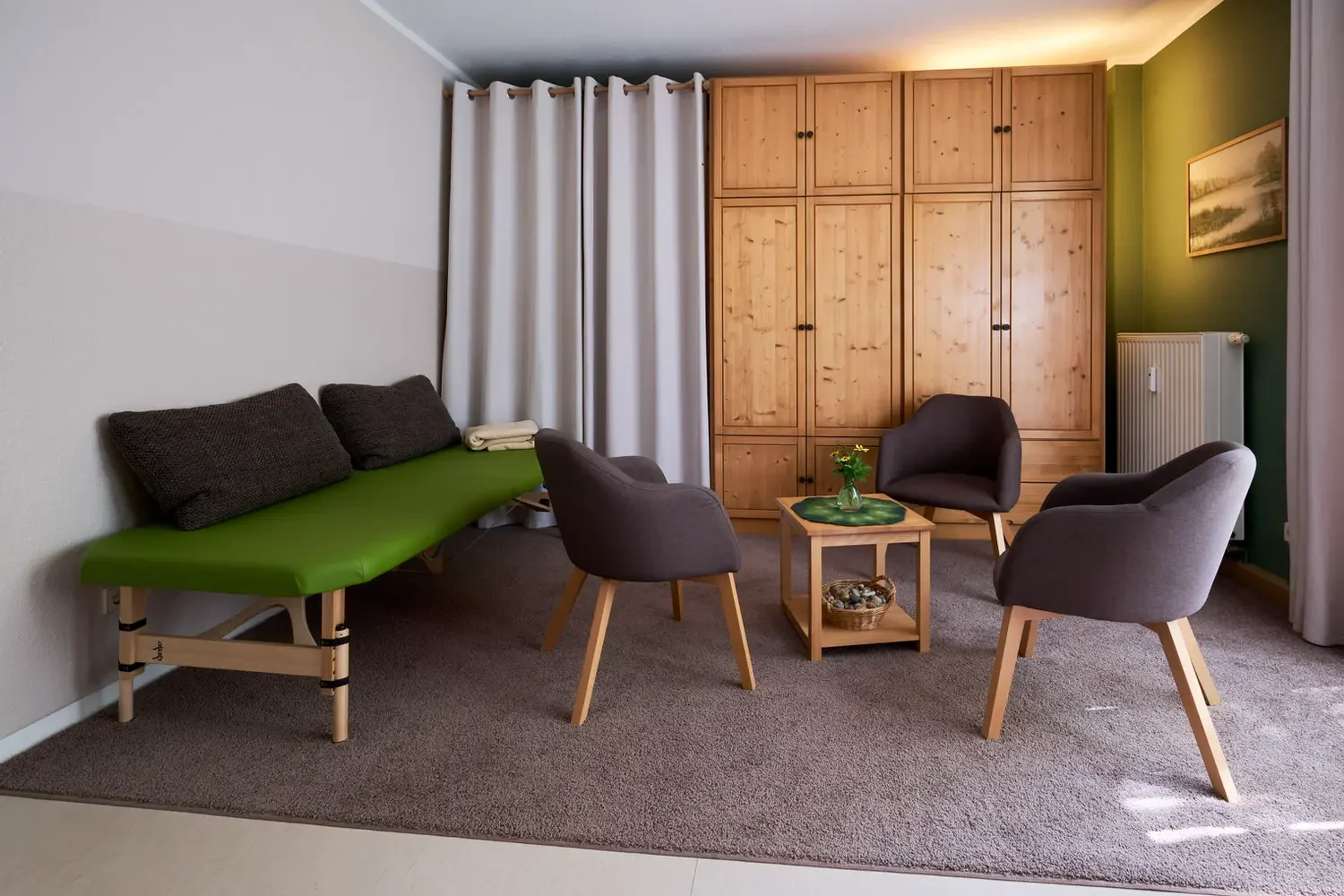 Gemütliches Wohnzimmer mit grünem Sofa, vier dunklen und hellgrauen Stühlen, einem kleinen Holztisch mit Blume und einer großen Holzkastennische. Weißer Vorhang und grüne Wand mit Bild dezenten Licht.