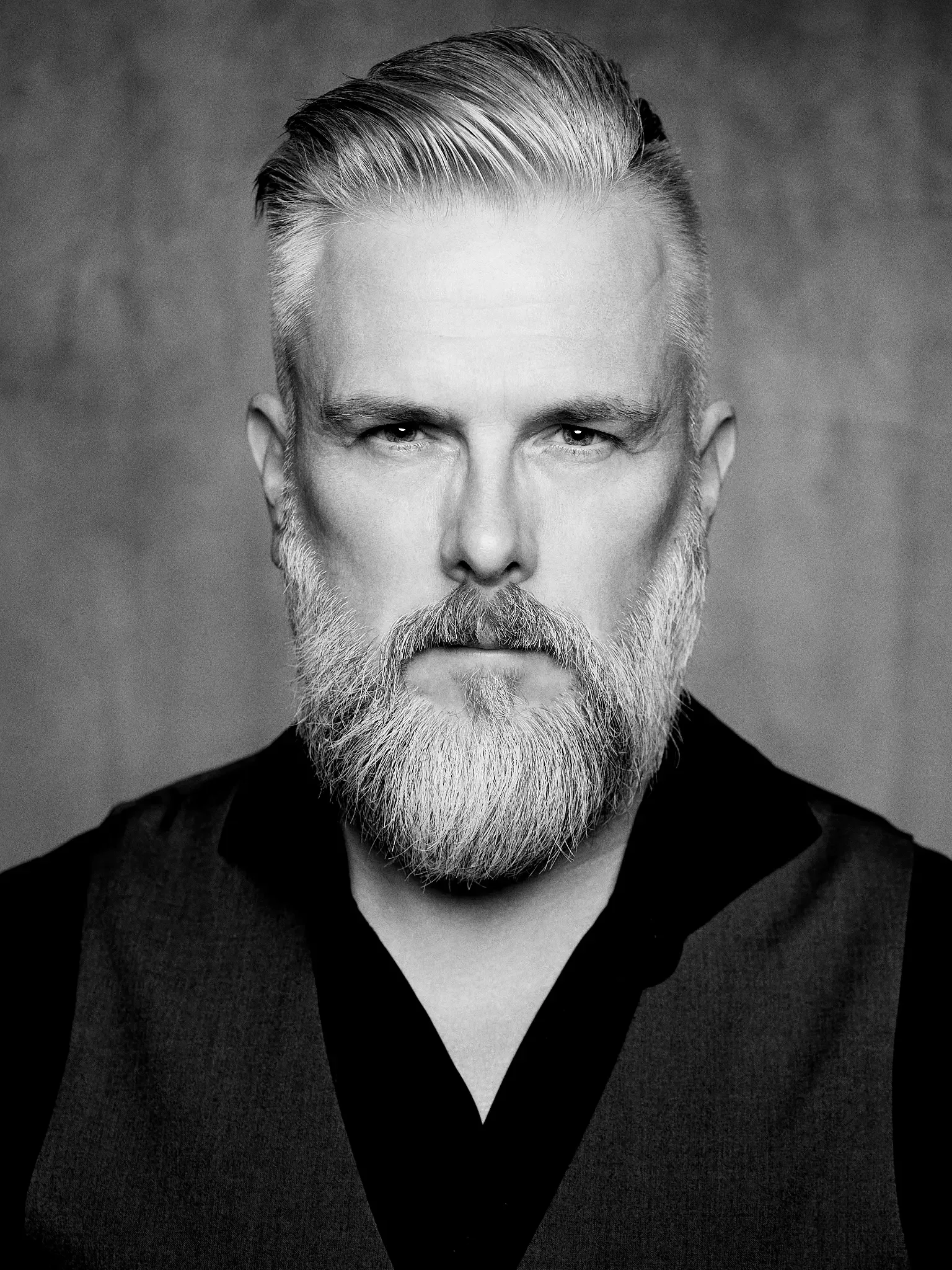 Ein schwarz-weiß Portrait eines Mannes mit einem gepflegten, grauen Bart und kurzen, nach hinten gestylten Haaren, trägt eine dunkle Weste und ein Hemd.