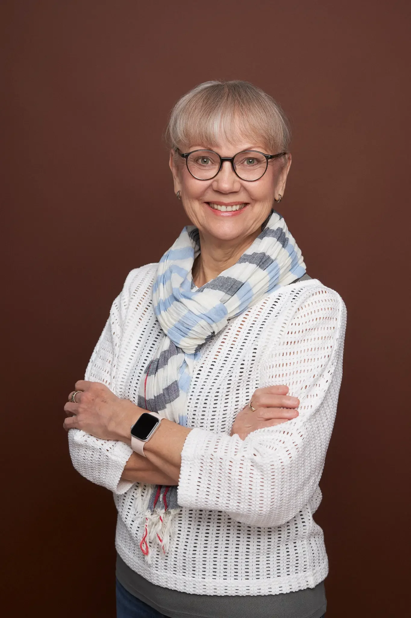 Eine lächelnde ältere Frau mit Brille, Kurzhaarfrisur, Weißem Pullover mit Lochmuster, blau-weiß gestreiftem Schal, Smartwatch am linken Handgelenk, vor braunem Hintergrund.