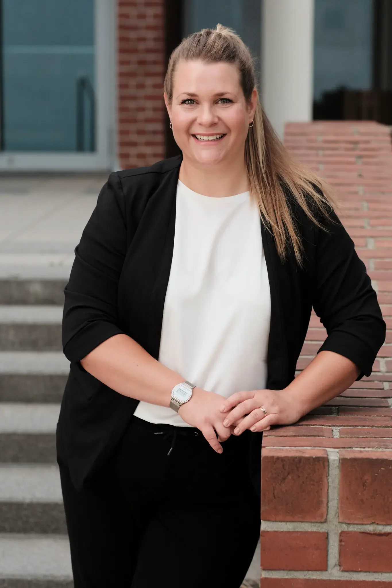 Eine lächelnde Frau mit braunem Haar, die eine schwarze Jacke, weiße Bluse und eine Armbanduhr trägt, steht draußen vor einer modernen Gebäude mit Ziegelmauer und Treppe.