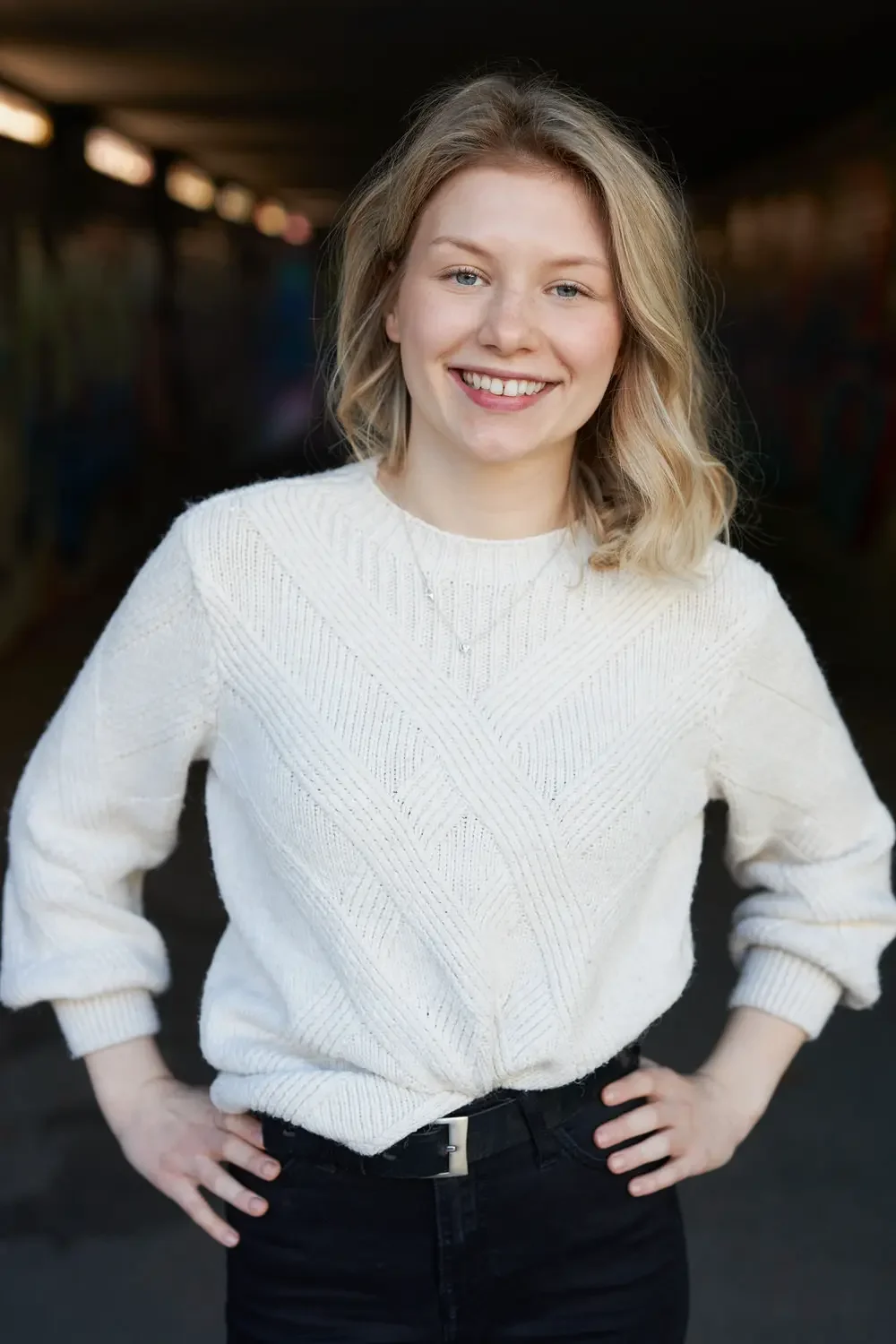 Junge Frau mit blonden Haaren, weißem Pullover und schwarzer Hose, lächelnd.