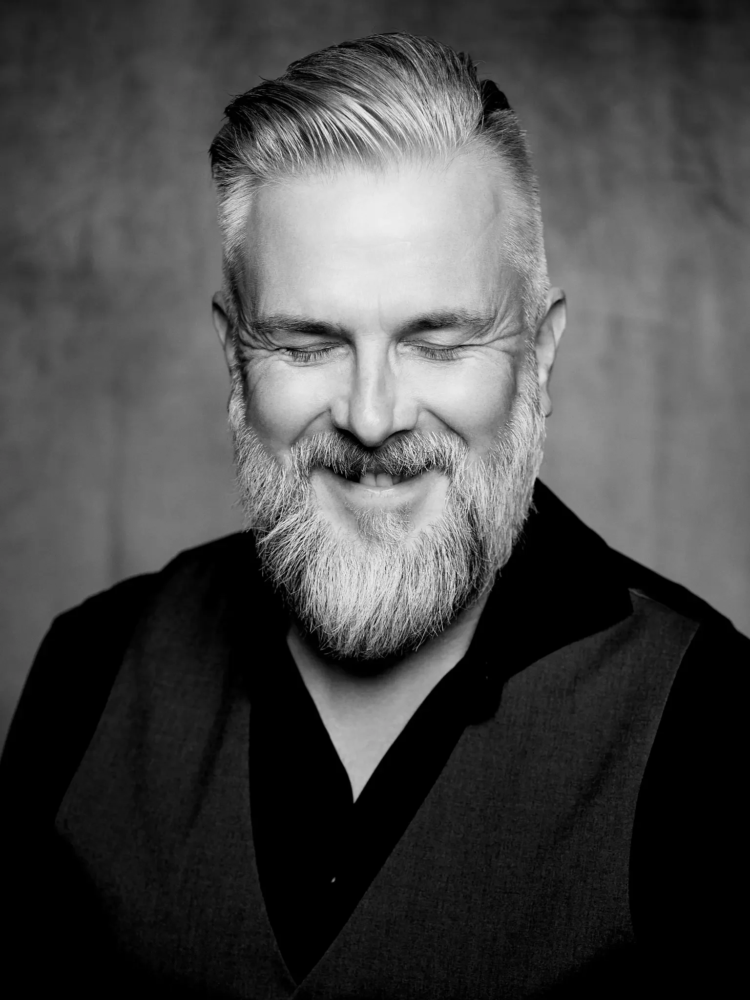 Schwarz-Weiß-Fotografie eines lachenden Mannes mit geschwätztem grauem Bart und gepflegtem Haar, trägt dunkle Kleidung, vor neutralem Hintergrund.