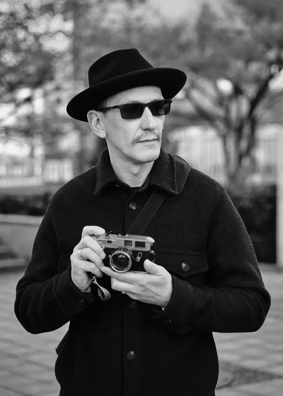 Schwarz-Weiß-Foto eines Mannes mit Hut, Sonnenbrille und Mantel, der eine Kamera in den Händen hält, im Hintergrund Bäume.