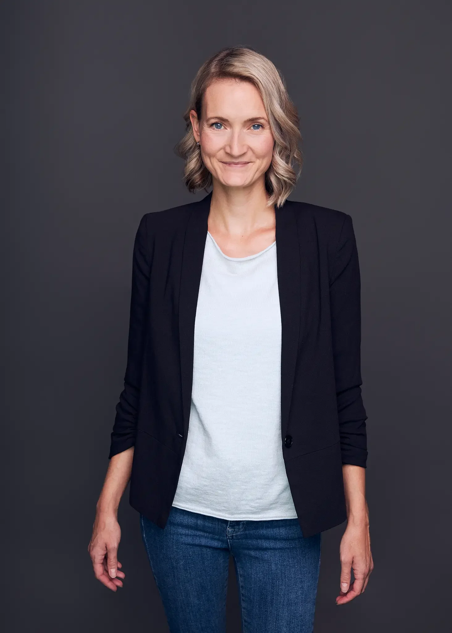 Eine Frau mit blonden, lockigen Haaren, die lächelnd in die Kamera schaut, trägt eine schwarze Blazer, ein weißes T-Shirt und blaue Jeans vor einem dunklen Hintergrund.