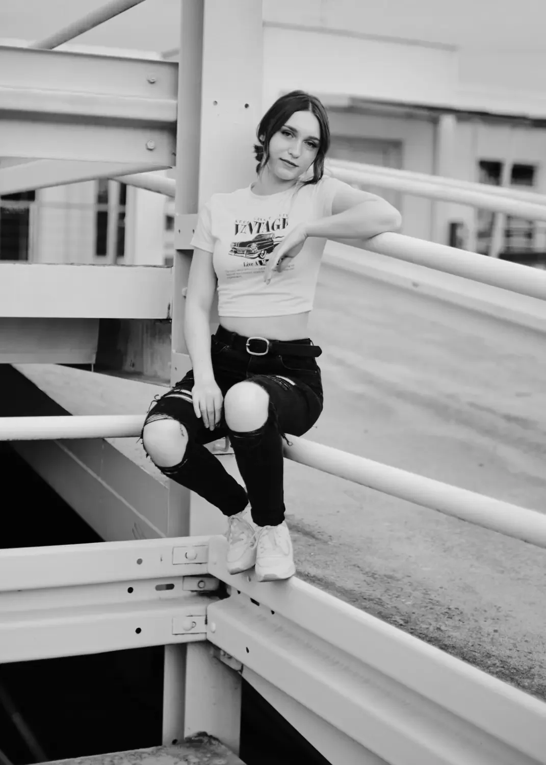 Schwarz-Weiß-Foto eines jungen Mädchens, das auf einem Parkhausgeländer sitzt, mit löchrigen Jeans und einem weißen T-Shirt, das einen alten Autoschriftzug zeigt, und Sneakers.