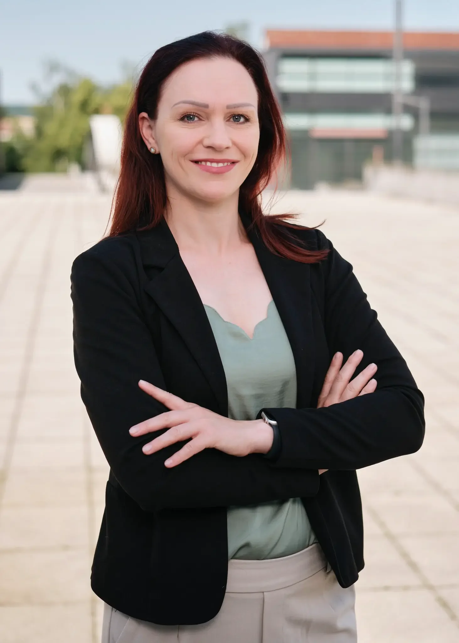 Porträt einer Frau mit roten Haaren, schwarzen Blazer, hellgrünem Oberteil und beige Hose, die auf einem Außenplatz steht, die Arme verschränkt und lächelt.