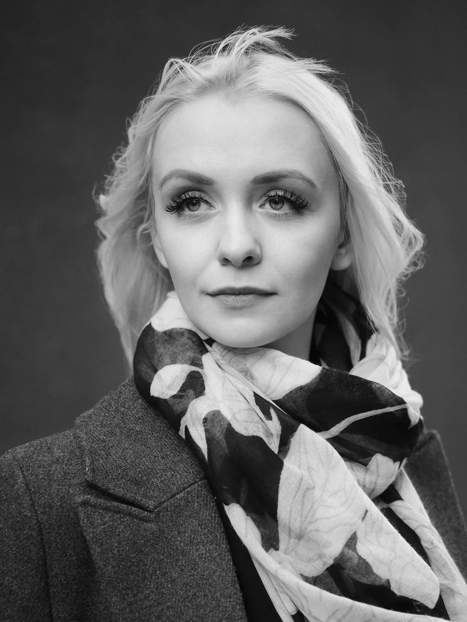 Schwarz-Weiß-Bild einer Frau mit blondem, lockigem Haar, trägt einen dunklen Mantel und ein farbiges Seidentuch. Blick nach links, gegen dunklen Hintergrund.