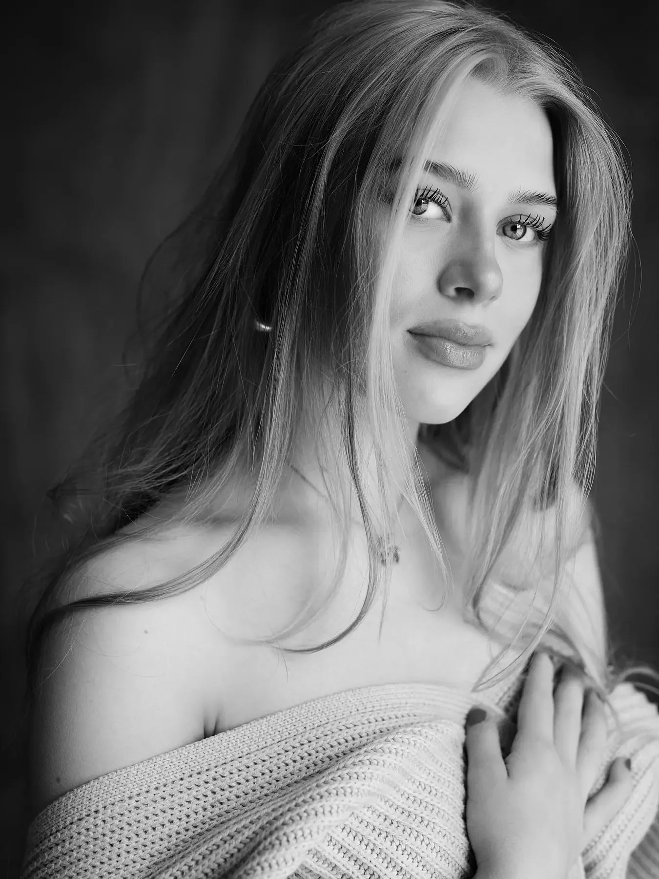 Schwarz-weiß-Porträt einer jungen Frau mit langen Haaren, die an einer Schulter angelehnt ist, mit einem dezenten Lächeln und Blick in die Kamera.
