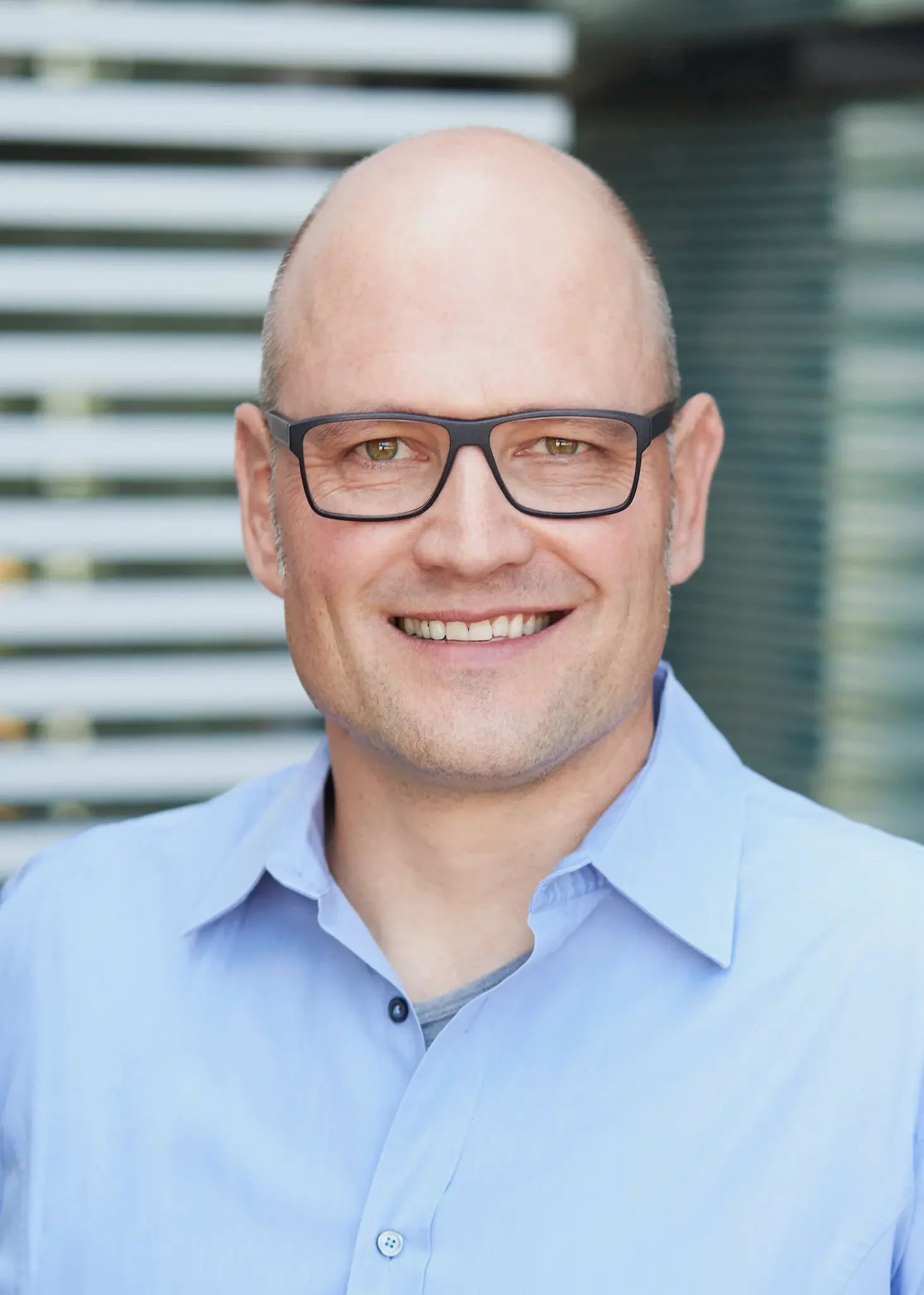 Ein lächelnder Mann mit Brille in einem blauen Hemd, im Hintergrund moderne Architektur.