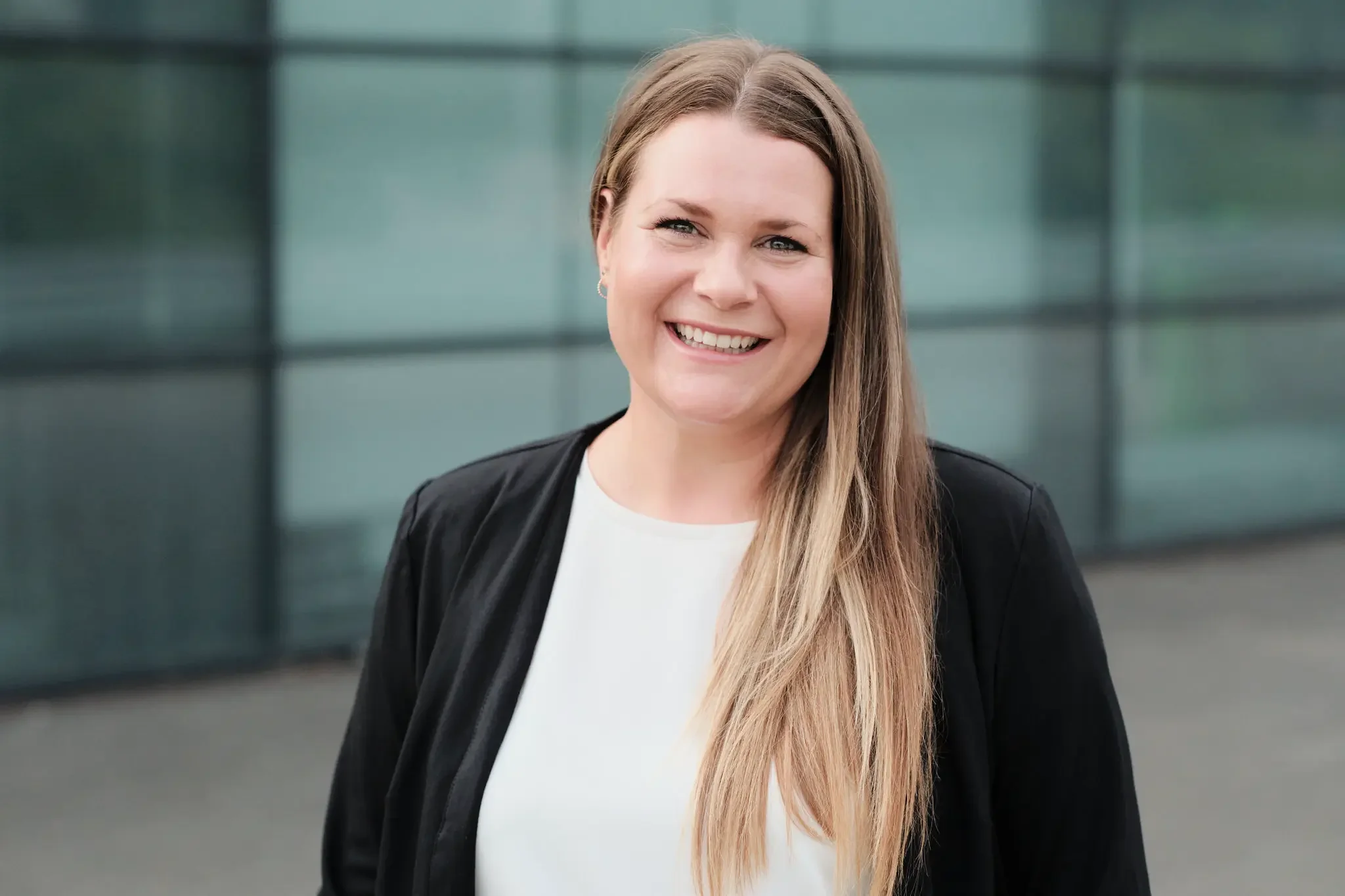 Lächelnde Frau mit braunem Haar, schwarzem Blazer und weißen Oberteil vor einem modernen Glasgebäude.