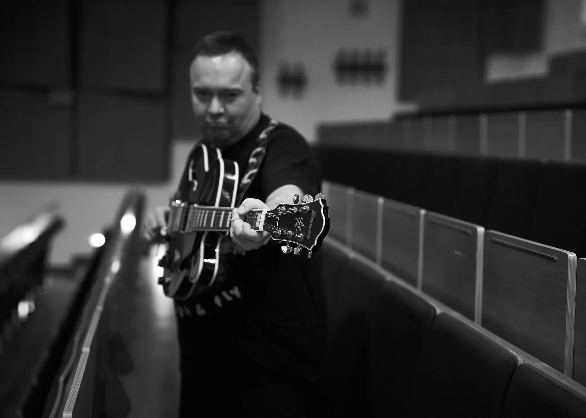 Ein Mann spielt Gitarre in einem Konzertsaal, schwarz-weiß Foto, Blick nach vorne, Sitzreihen im Hintergrund.
