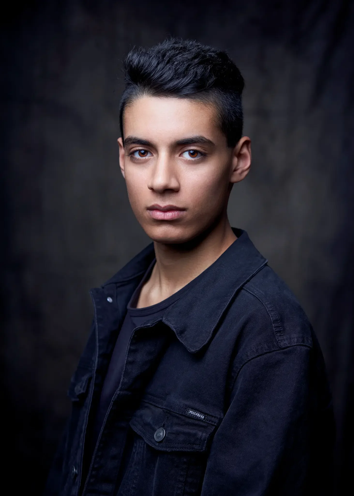 Porträt eines jungen Mannes mit dunklem Haar, blauen Augen, trägt eine dunkle Jacke vor einem dunklen, unscharfen Hintergrund.