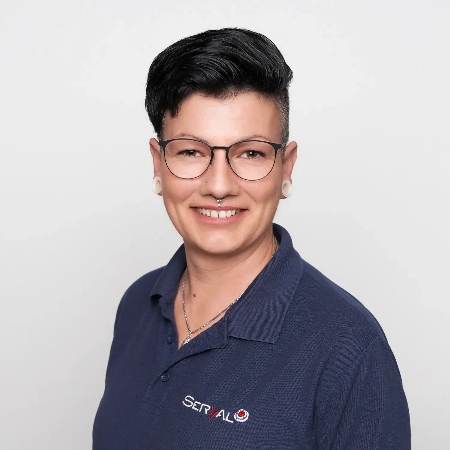 Porträt einer Person mit kurzen schwarzen Haaren, Brille, Piercings, im blauen Polo mit Firmenlogo, lächelnd vor weißem Hintergrund.