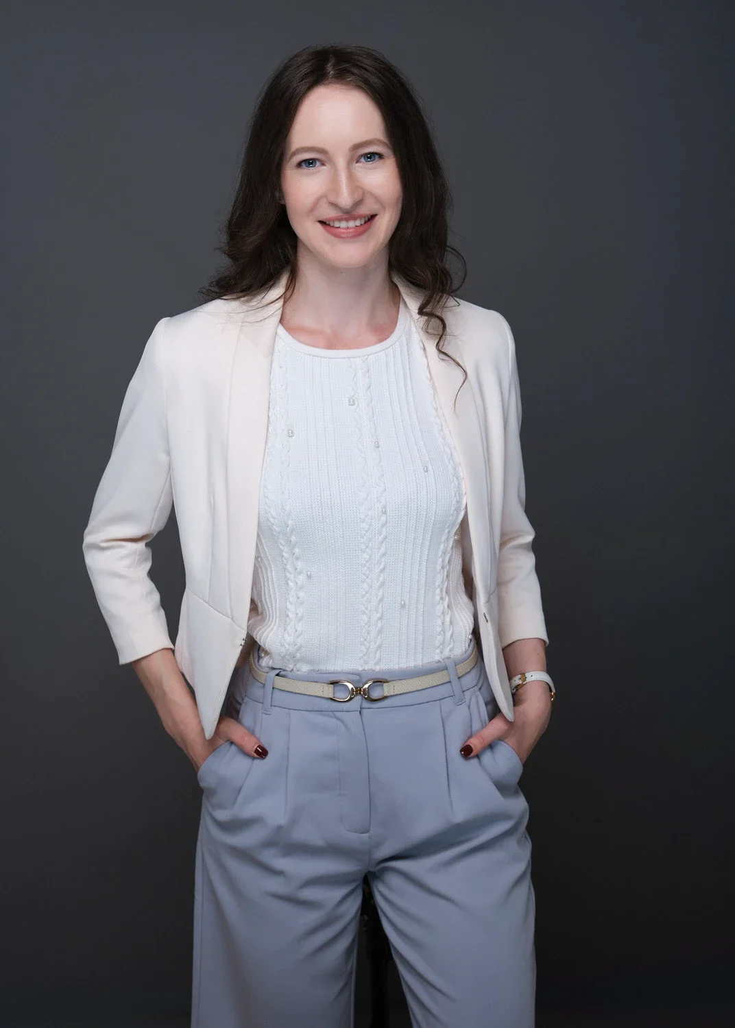 Eine Frau mit dunklen, lockigen Haaren trägt eine weiße Jacke, eine weiße Strickbluse und hellgraue Hosen. Sie steht vor einem dunklen Hintergrund und lächelt in die Kamera.