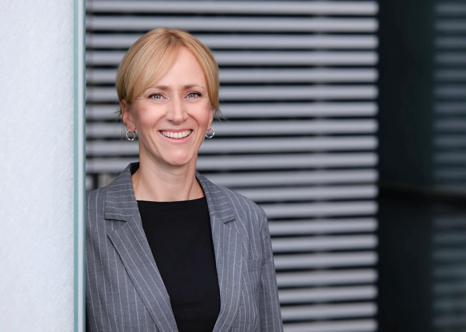 Ein Lächelnde Frau in einem grauen Businessanzug steht an einer Wand mit horizontalen Linien im Hintergrund.