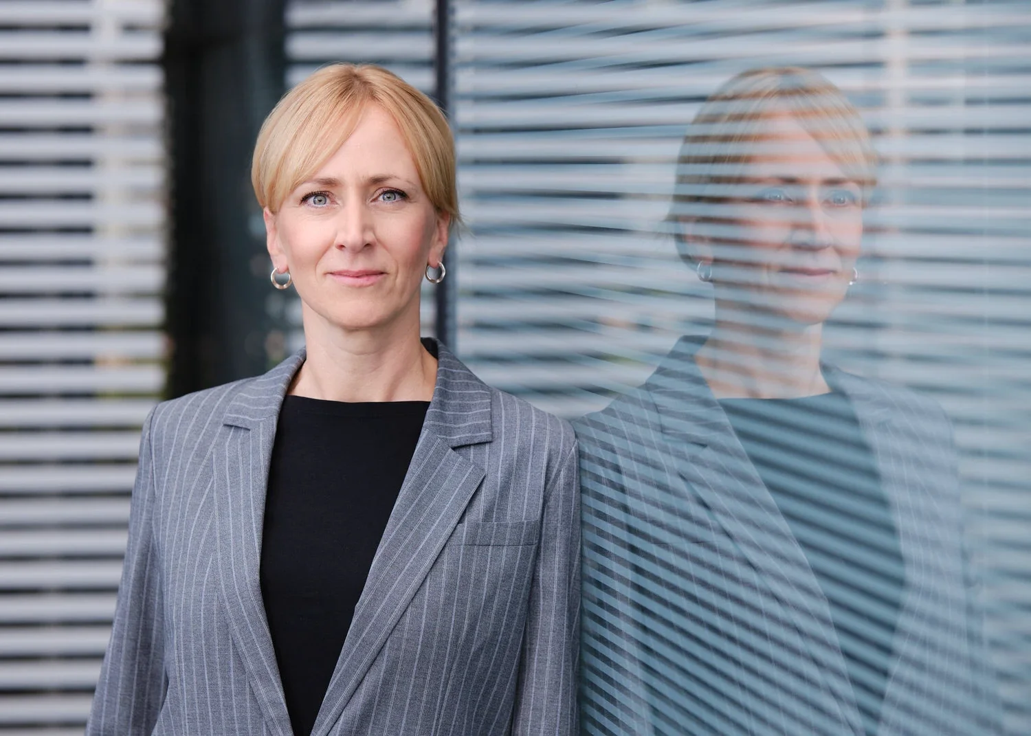 Eine blonde Frau trägt einen grauen Anzug und steht vor einem Fenster, an dem ihre Reflexion sichtbar ist.