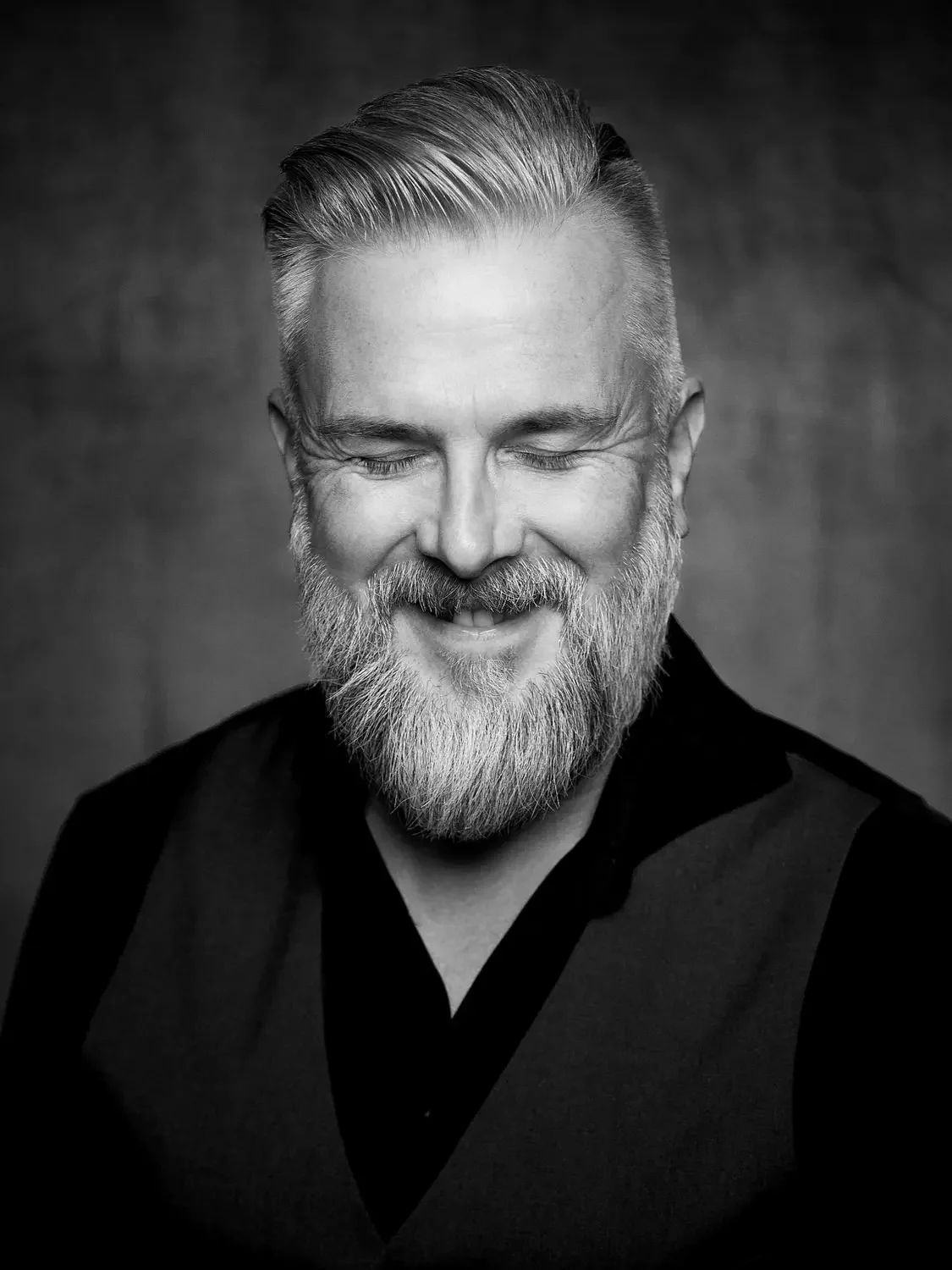 Schwarz-Weiß-Porträt eines lachenden, älteren Mannes mit grauem, gepflegtem Bart und kurzen, zurückgekämmten Haaren, trägt dunkle Kleidung.