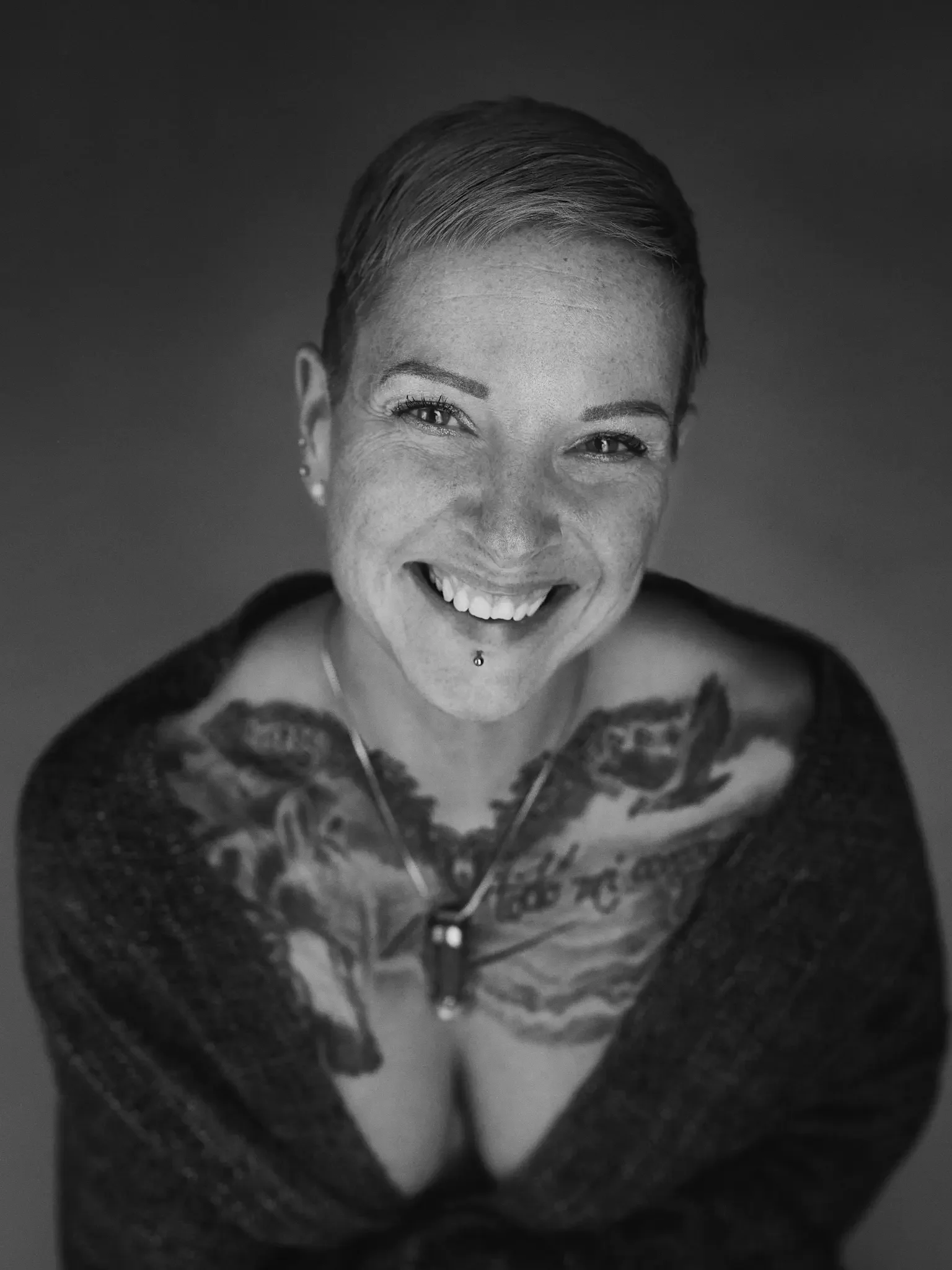 Schwarz-weiß Portrait einer lachenden Frau mit kurzen Haaren, Tattoos auf den Schultern und einem Piercing auf der Unterlippe, trägt dunklen Pullover und eine Halskette.