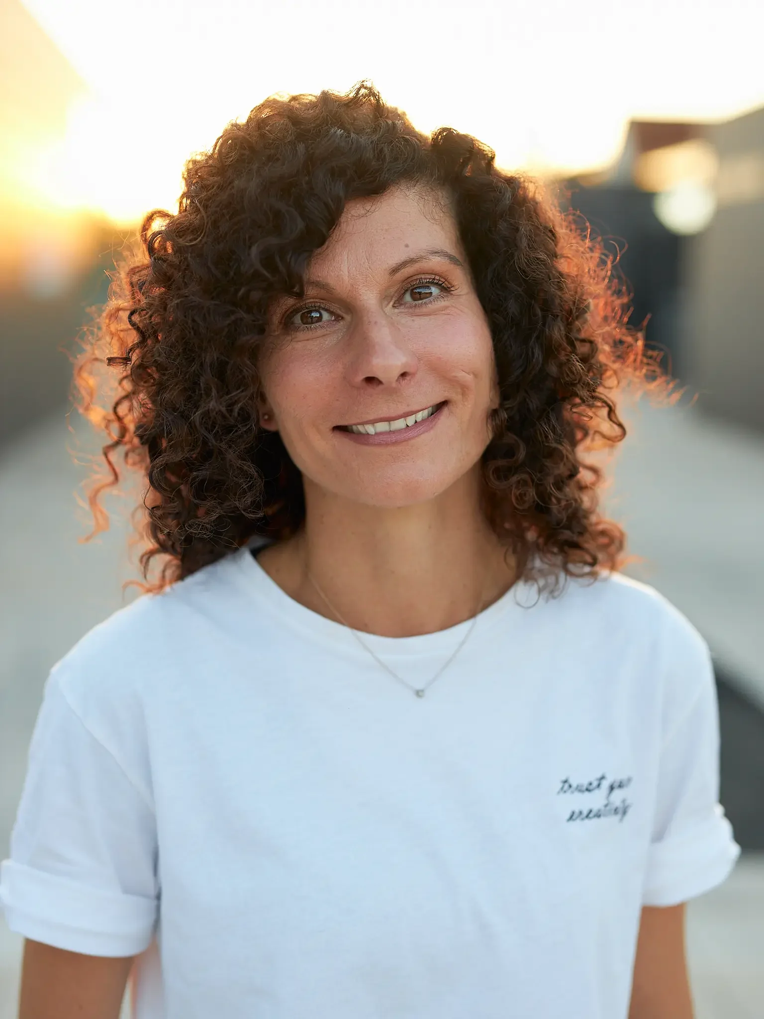 Porträt einer lachenden Frau mit lockigem Haar, Sonnenuntergang im Hintergrund, weißes T-Shirt.