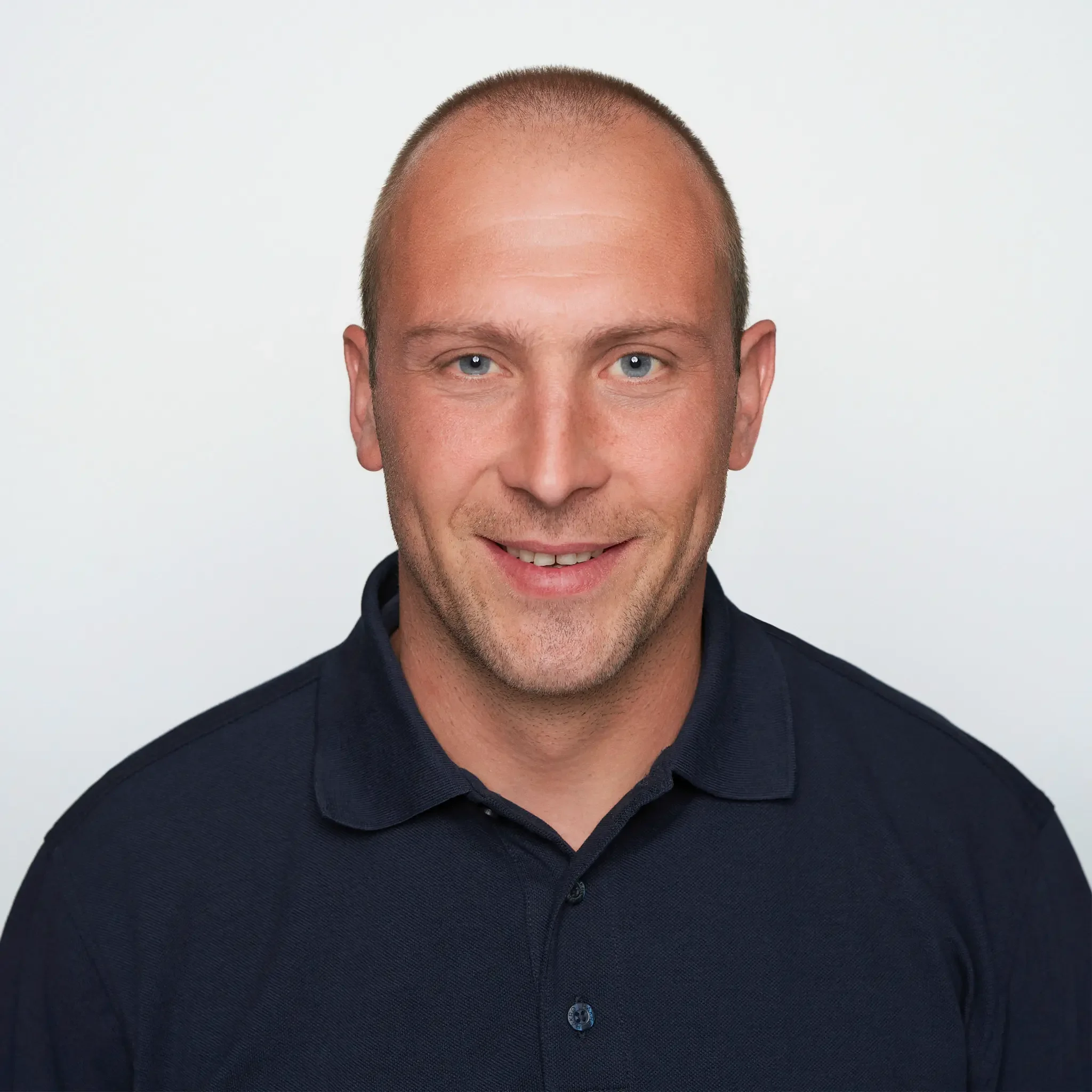 Porträt eines lächelnden Mannes mit kurzen Haaren, hellblauen Augen und dunklem Hemd auf weißem Hintergrund.