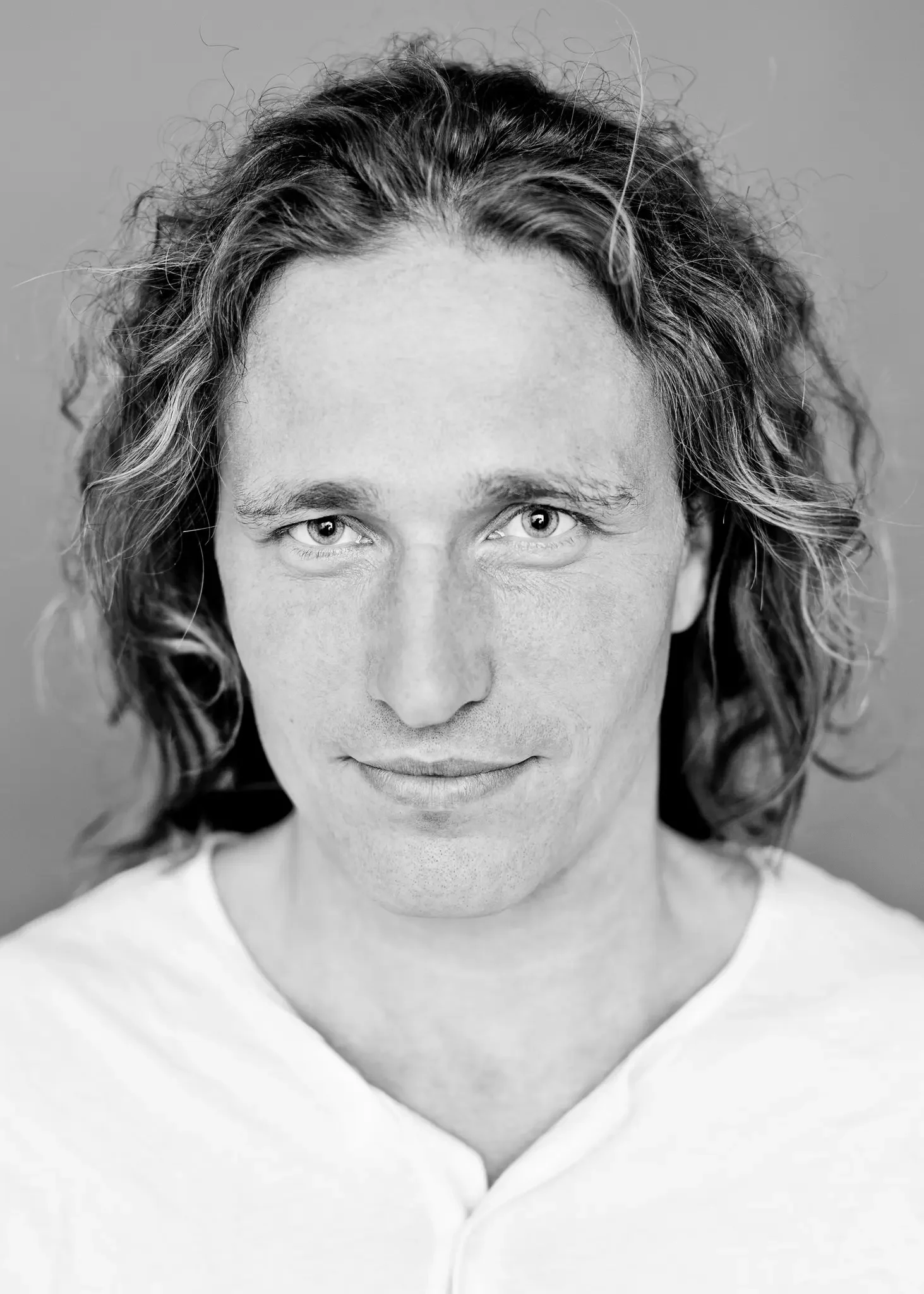Schwarz-weiß Portrait eines Mannes mit langen, lockigen Haaren, mittelalter Haut, und eines weißen T-Shirts, lächelnd, vor hellem Hintergrund.