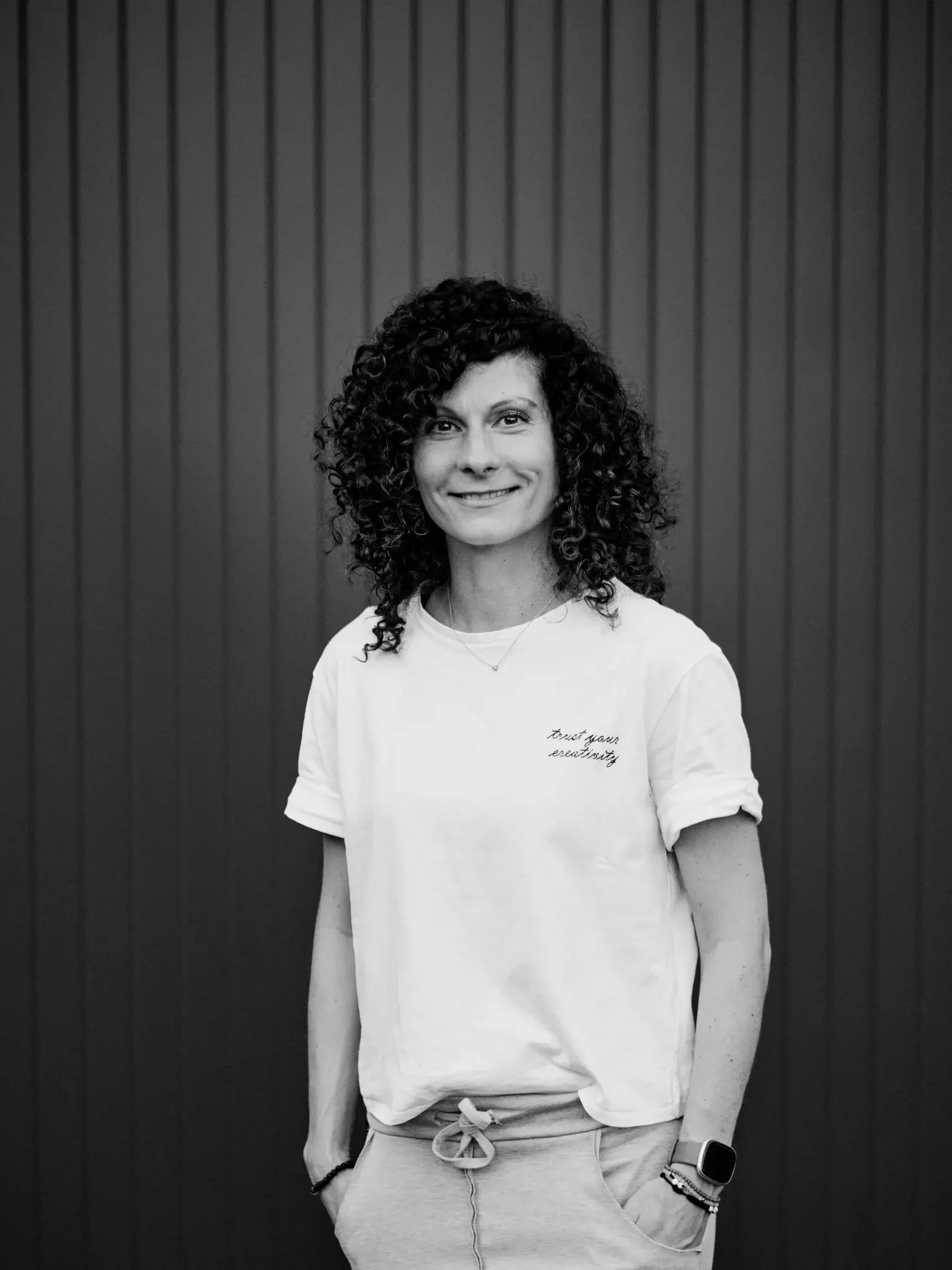 Schwarz-weiße Portraitaufnahme einer lächelnden Frau mit lockigem Haar, die casual Kleidung trägt, vor einer vertikal gestalteten dunklen Wand.