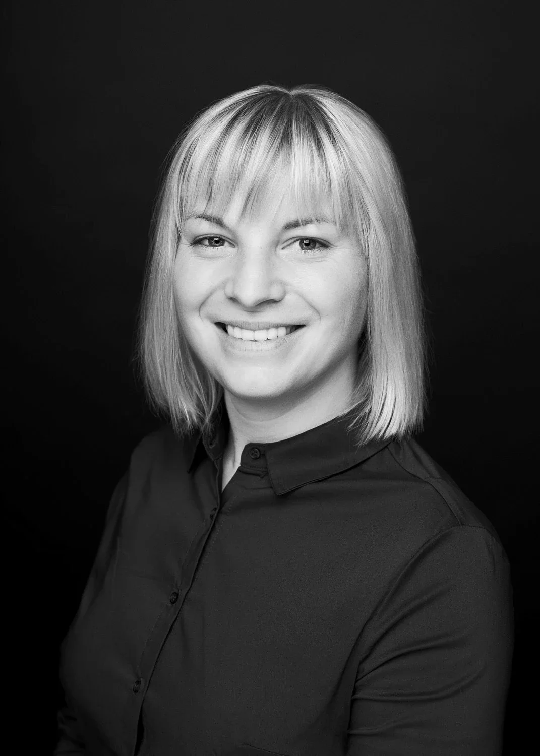 Schwarz-Weiß-Foto einer lachenden Frau mit Schulterlänge blondem Haar, getragen einem dunklen Hemd, vor dunkelhäutigem Hintergrund.