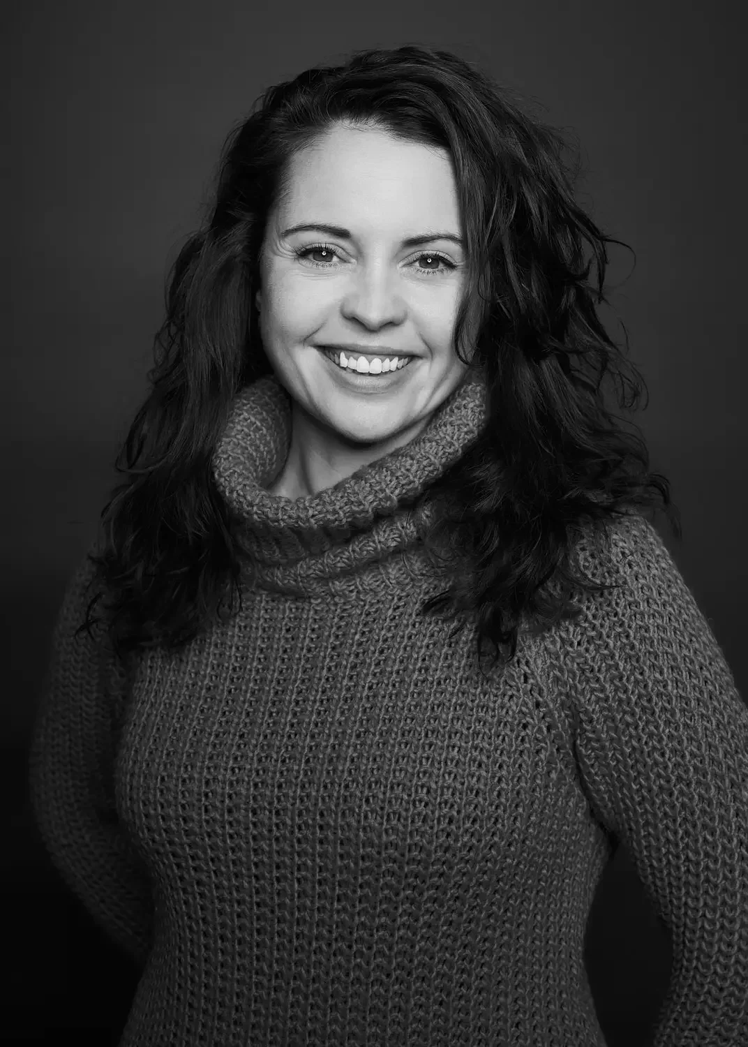 Schwarze und weiße Porträtaufnahme einer lächelnden Frau mit lockigen Haaren, die Kabelstrickpullover trägt.