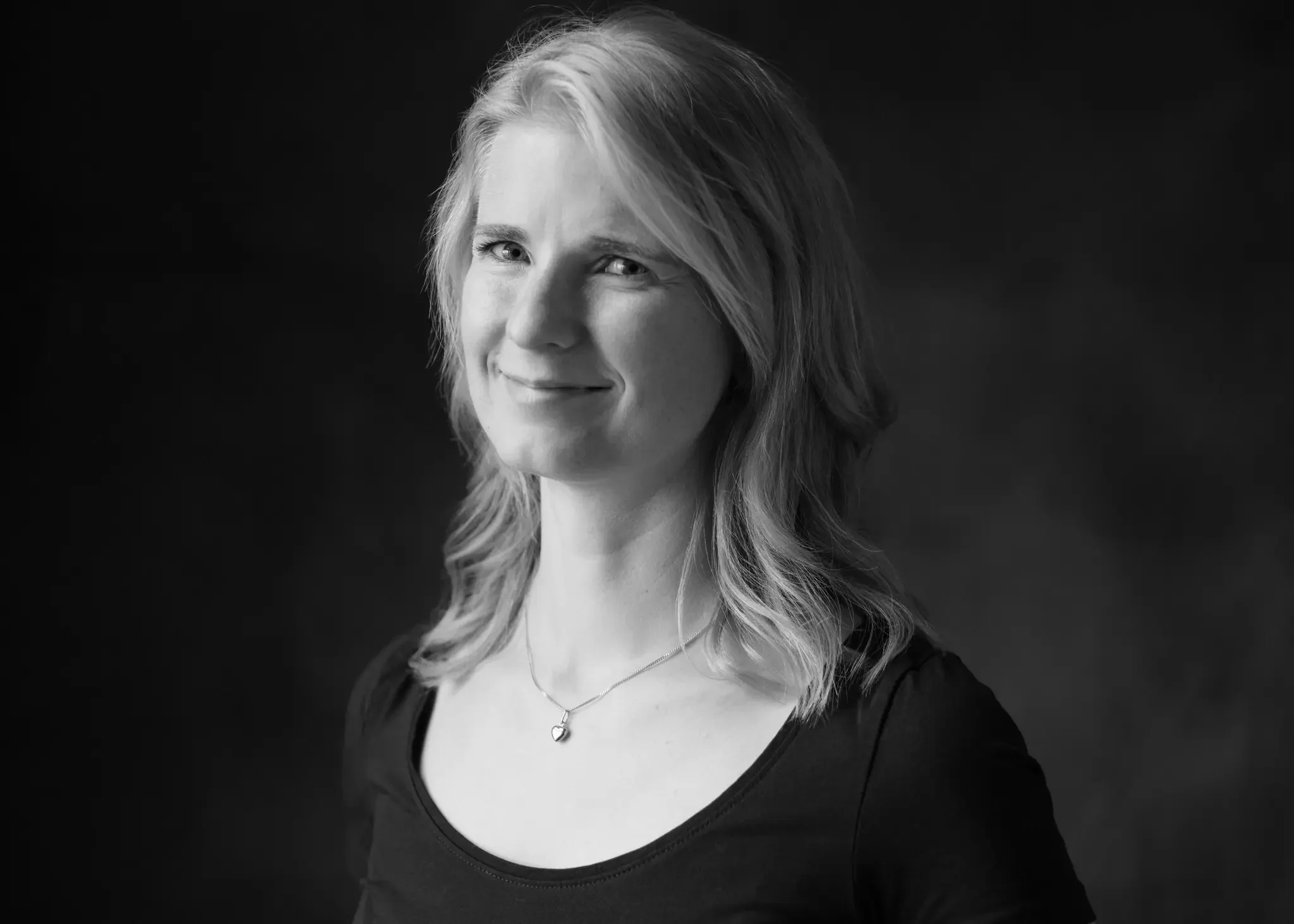 Schwarz-Weiß-Foto eines lächelnden Frau mit Schulterlänge, mittellangem, gewelltem Haar, trägt ein dunkles Oberteil und eine Halskette mit Anhänger.