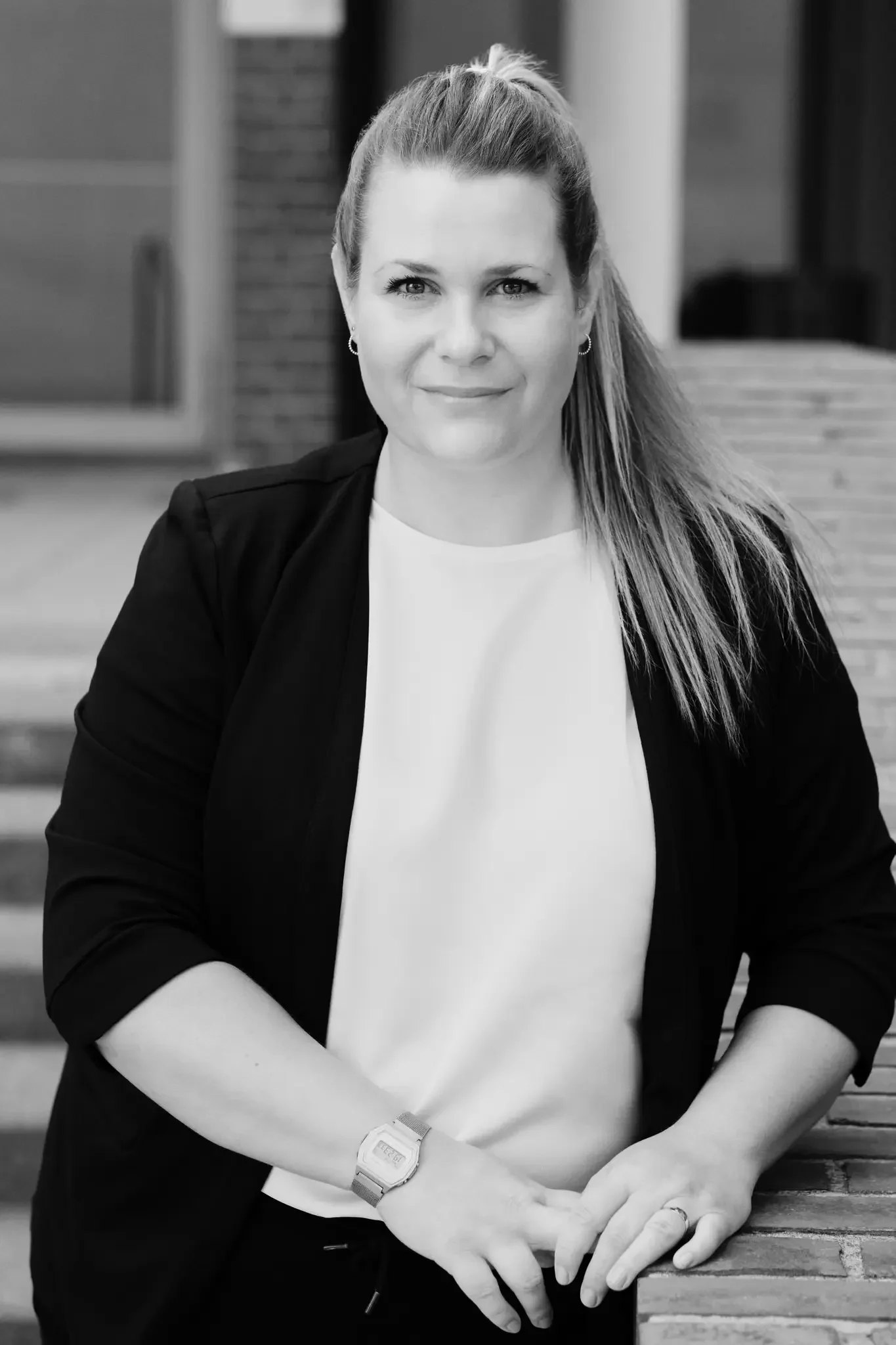 Schwarz-weiß Portrait einer Frau mit langem Haar, die eine weiße Bluse und einen schwarzen Blazer trägt, vor einem Hintergrund aus Ziegeln und Holz. Sie steht an einem Tisch und schaut in die Kamera.