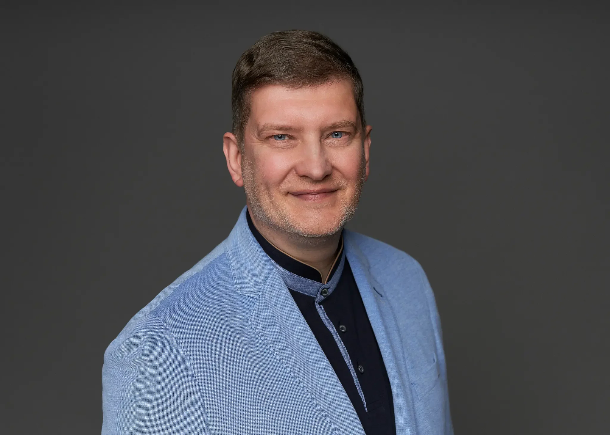 Porträt eines Mannes mit blauen Augen, braunen Haaren und einem leichten Bart, trägt eine hellblaue Jacke und ein dunkles Hemd, vor grauem Hintergrund.