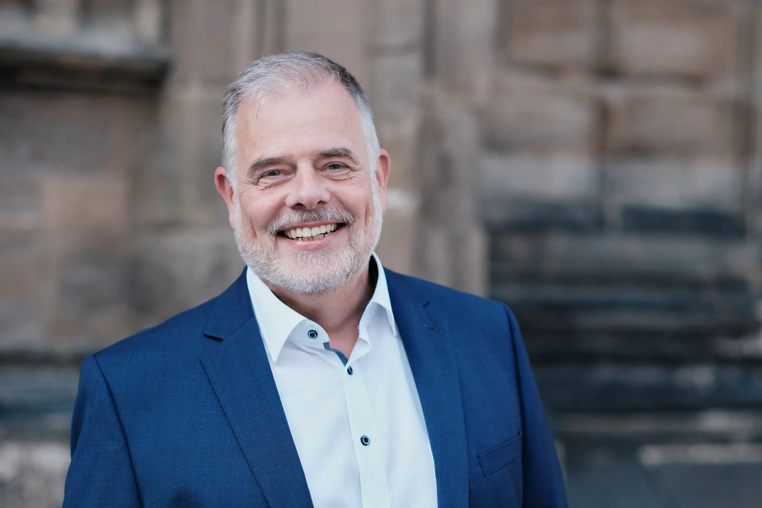 Ein älterer Mann mit grauem Haar und Bart lächelt, trägt einen dunkelblauen Anzug und ein weißes Hemd, im Hintergrund eine Steinmauer.