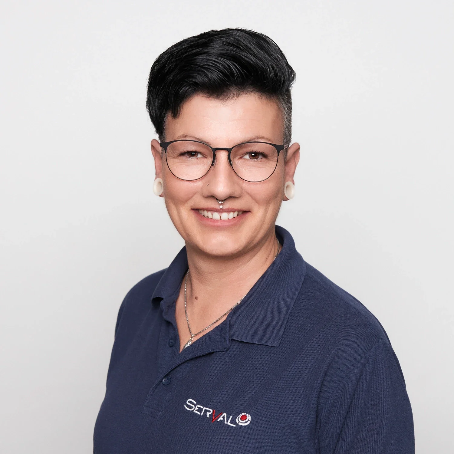 Porträt einer Person mit kurzen schwarzen Haaren, Brille, Nasenpiercing, großen Ohrhörern, im blauen Polo mit Logo auf der Brust, lächelnd vor weißem Hintergrund.