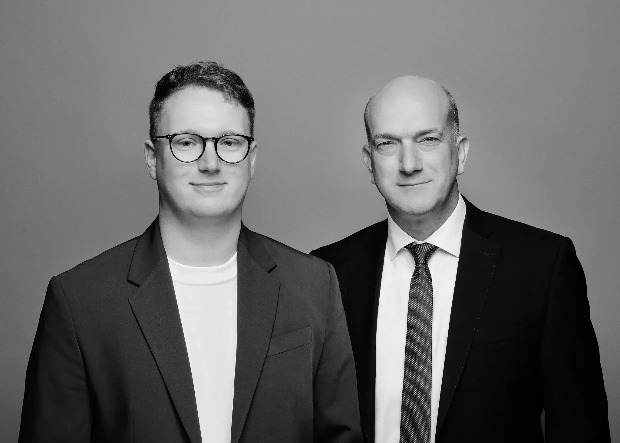 Zwei Männer in formeller Kleidung, einer trägt Brille, stehen nebeneinander vor einem einfarbigen Hintergrund.