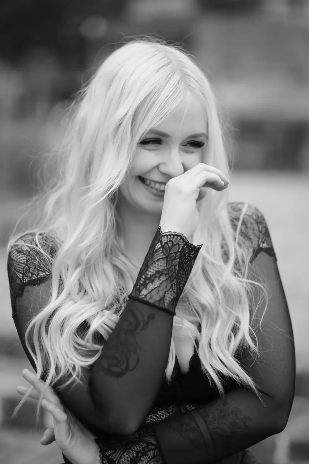 Lächelnde Frau mit langen, lockigen, blonden Haaren in dunklem Spitzenkleid, lachend und die Hand vor Mund haltend, im Freien.