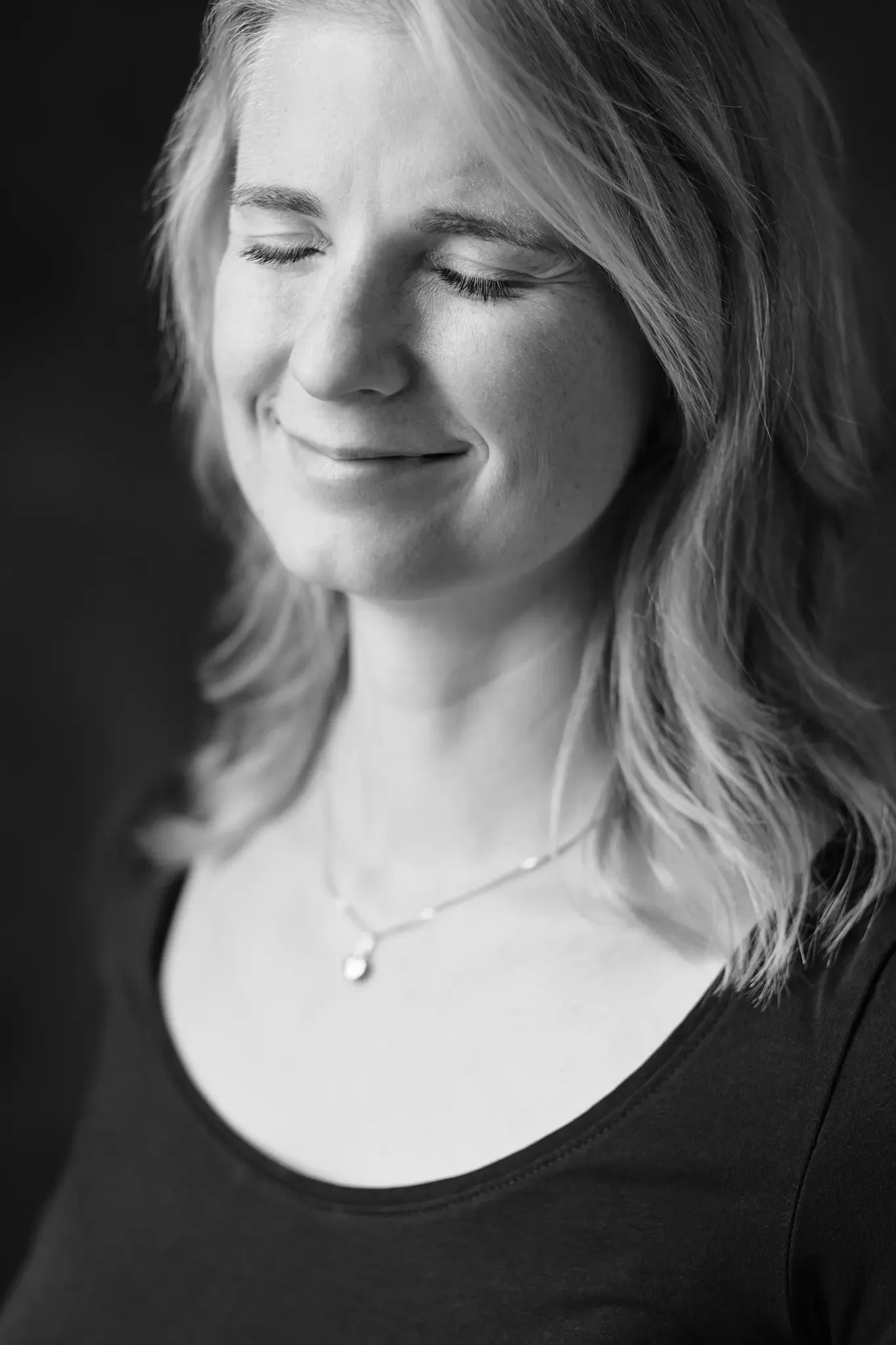 Schwarz-weiß-Fotografie eines lächelnden Frau mit geschlossenen Augen, mittellange wellige Haare und zarte Halskette, dunkles Oberteil.