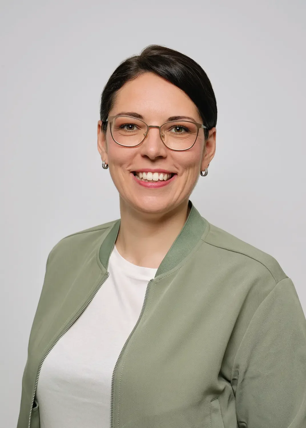 Porträt einer lächelnden Frau mit Kurzhaarfrisur, Brille und Echtschmuck, trägt einen grünen Blouson und ein weißes Shirt vor hellem Hintergrund.