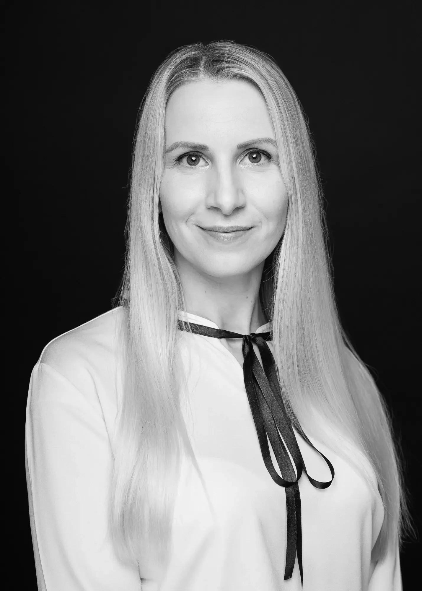 Schwarz-Weiß-Fotografie einer lächelnden Frau mit langen, glatten Haaren, trägt eine weiße Bluse mit schwarzer Schleife gegen schwarzem Hintergrund.