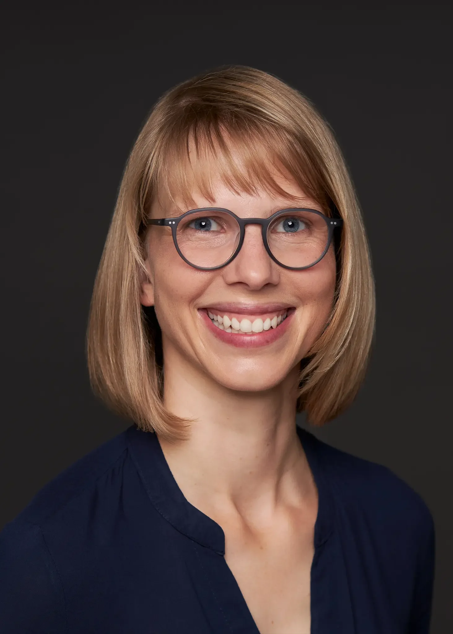 Porträt einer Frau mit Brille und Lächeln vor dunklem Hintergrund