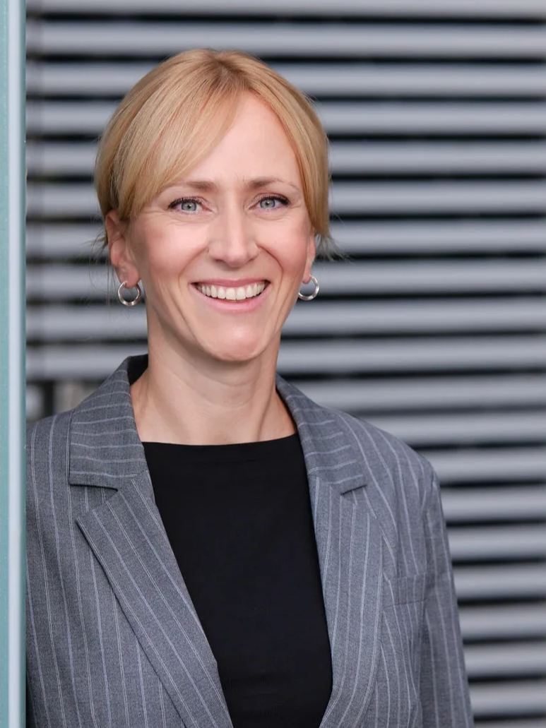 Porträt einer lächelnden Frau mit kurzem blondem Haar, schwarze Bluse und grau gestreifter Blazer, im Hintergrund horizontal gestreifte Wand.