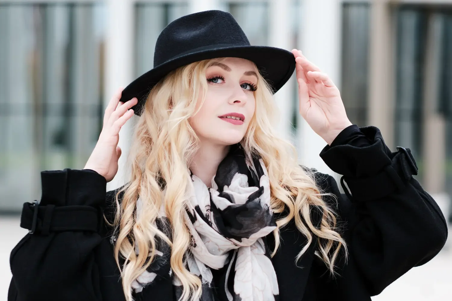 Eine junge Frau mit lang, lockigem blonden Haar trägt einen schwarzen Hut, einen schwarzen Mantel und ein helles Halstuch mit schwarz-weißen Mustern, posiert vor einem modernen Hintergrund.