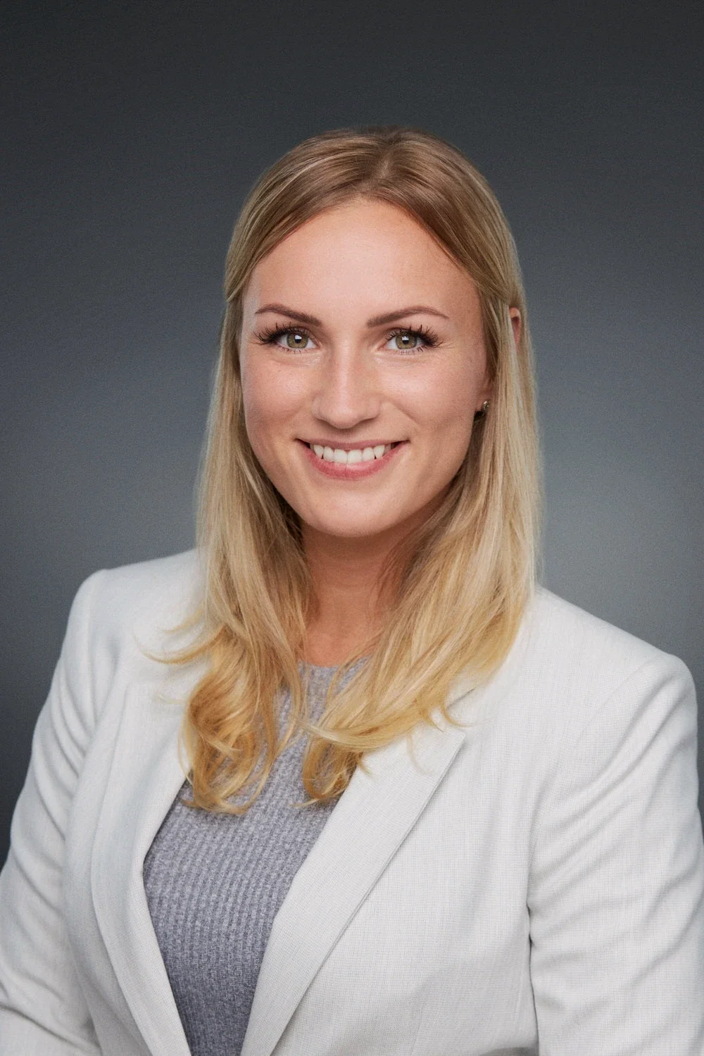 Porträt einer lächelnden Frau mit blonden Haaren, weißem Blazer und grauem Oberteil vor dunklem Hintergrund.