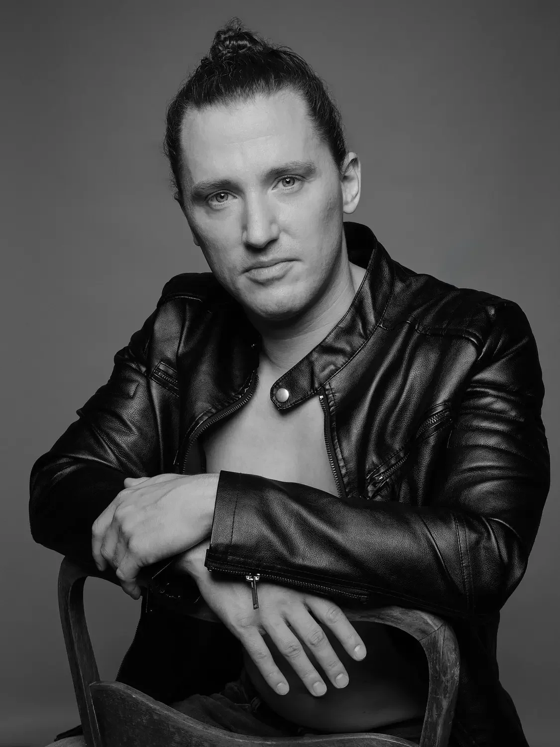 Schwarz-Weiß-Fotografie eines jungen Mannes mit dunklen Haaren in einem Dutt, der einen schwarzen Lederjacke trägt, mit einer nachdenklichen Ausstrahlung, sitzend auf einem Stuhl.