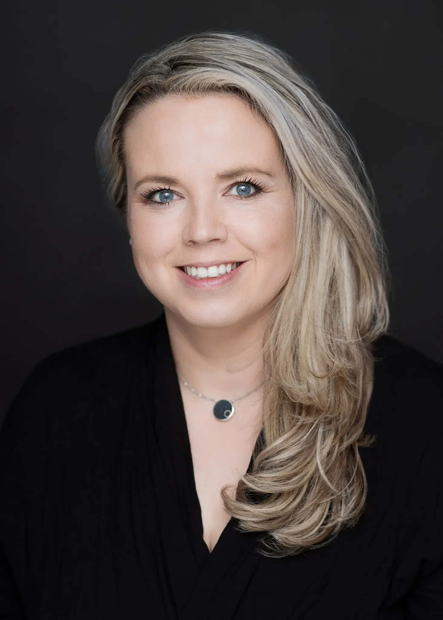 Porträt einer lächelnden Frau mit langen, blonden Haaren und blauen Augen, getragen auf einem schwarzen Hintergrund.