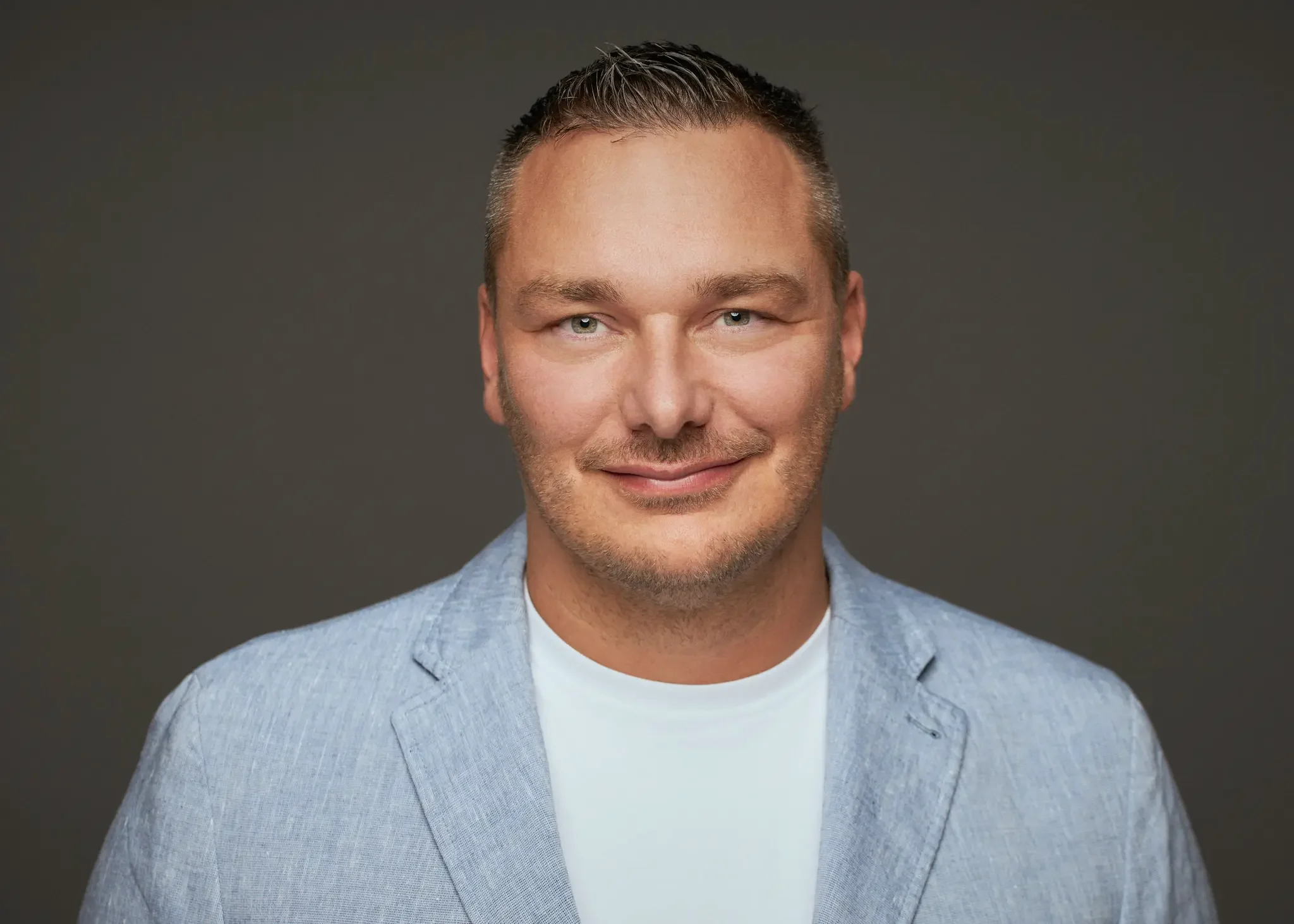 Mann mit kurzen hellbraunen Haaren, blauer Jacke und weißem T-Shirt vor grauem Hintergrund.
