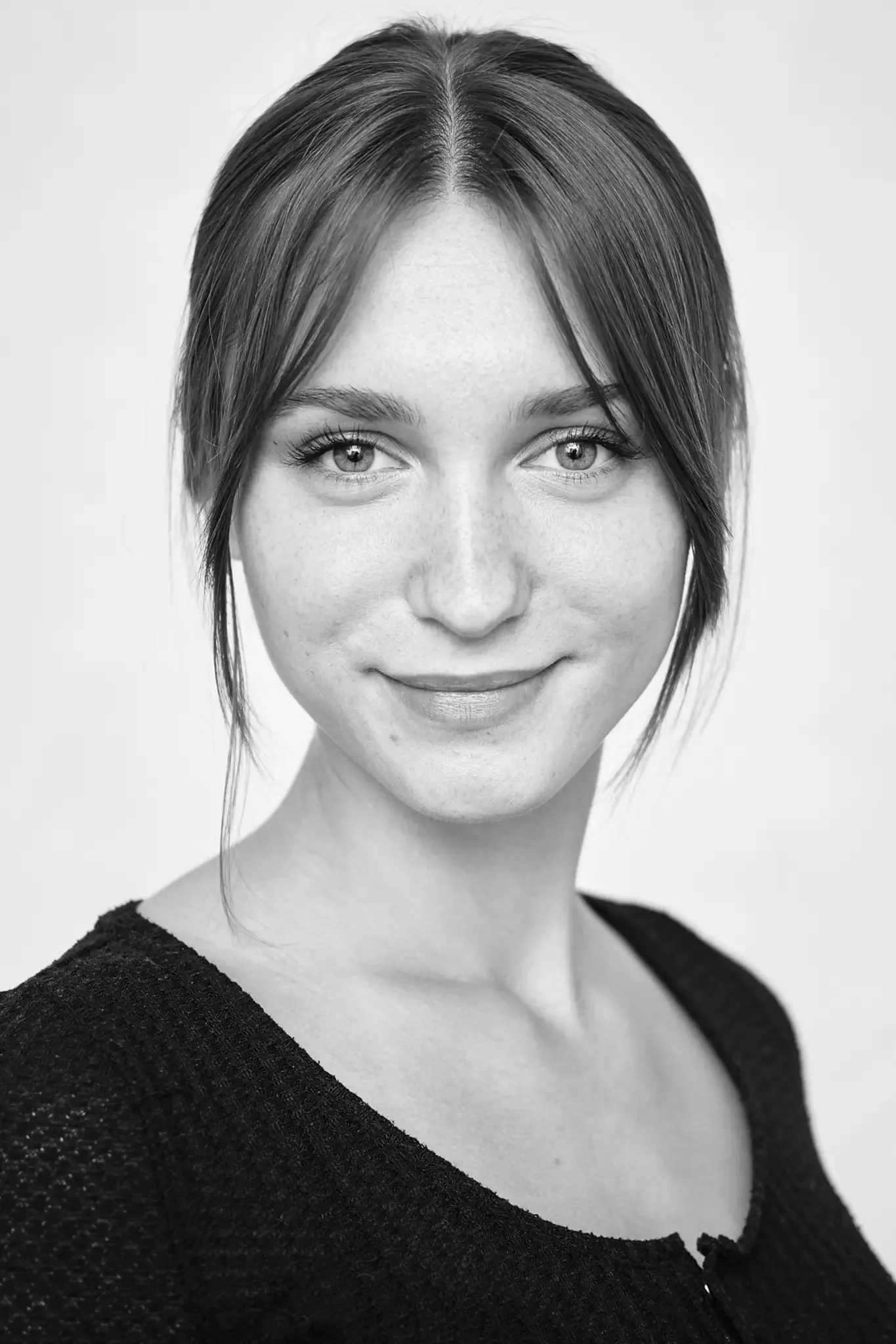 Schwarz-Weiß-Porträt einer jungen Frau mit kurzen Haaren, die leicht nach oben gestylt sind, trägt ein dunkles Oberteil, lächelt leicht und hat helle Augen.