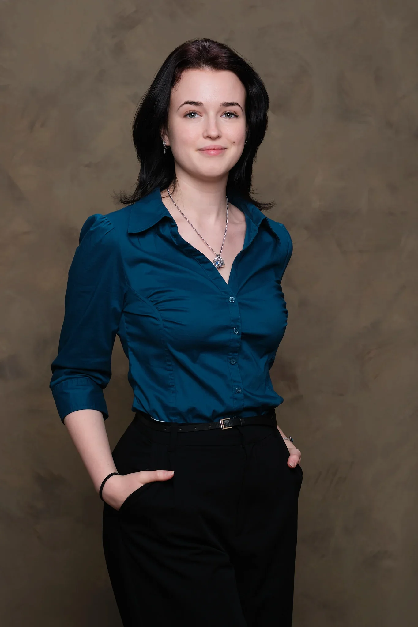 Junge Frau mit schwarzen Haaren in blauer Bluse und schwarzen Hosen vor braunem Hintergrund.