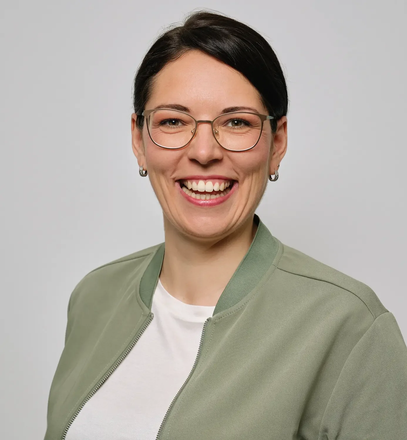 Lächelnde Frau mit Kurzhaarfrisur, Brille und grüner Jacke vor einem grauen Hintergrund.