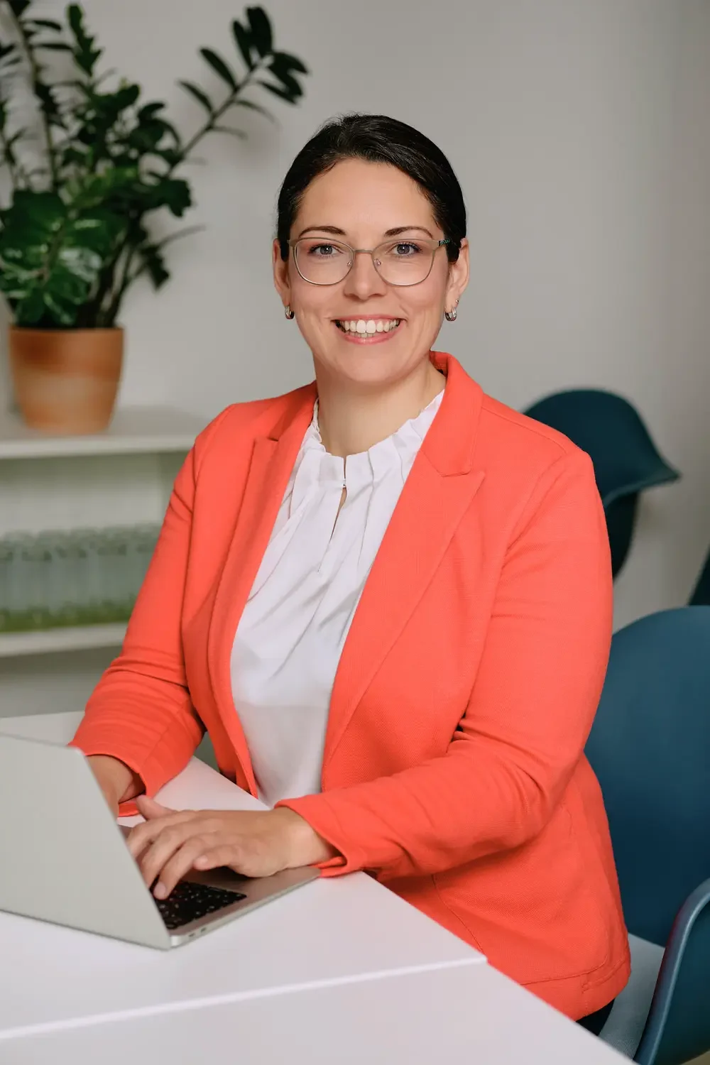 Eine lächelnde Frau mit dunklen Haaren, Brille und Ohrringen, trägt eine orange Jacke und eine weiße Bluse, sitzt an einem weißen Tisch mit einem Laptop; im Hintergrund ein grüner Pflanze in Tontopf und ein Stuhl.